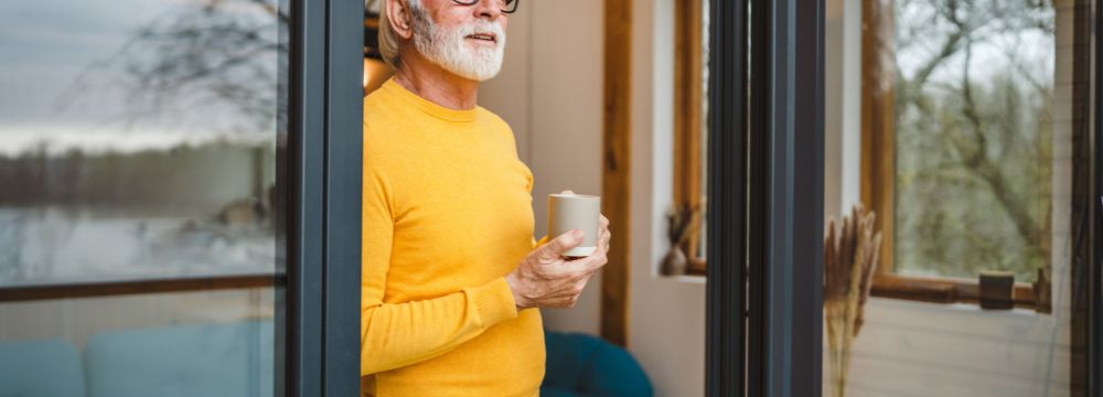 Older man looking out open sliding door window with coffee in hands