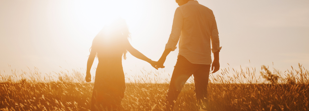 Male and female couple walking in sunny field during sunset holding hands 